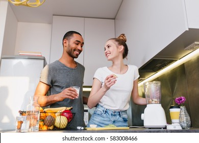 Couple In Home Clothes In Kitchen Talking With Happy Faces. Pair Have A Chat And Cooking A Meal At The Same Time. Warm Relationship, Healthy Eating, Smile On Faces.