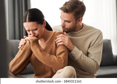 Couple at home. Handsome young man is calming his upset crying wife while holding her shoulders and apologizes. People, relationship difficulties, conflict and family concept