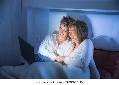 Couple At Home Watching A Movie On The Computer. Young Lgbt Couple Of Two Women Watching A Film In Bed. They Are In The Bedroom Sitting Together And Laughing.
