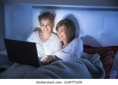 Couple At Home Watching A Movie On The Computer. Young Lgbt Couple Of Two Women Watching A Film In Bed. They Are In The Bedroom Sitting Together And Laughing.