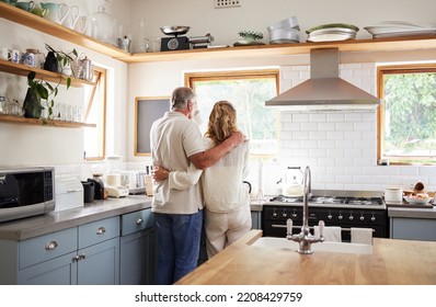 Couple Hug, Morning Coffee And Senior People Having Breakfast In The Kitchen While Looking At Outdoor Nature. Elderly Man And Woman Hugging With Tea Drink In House With Love Together From Behind