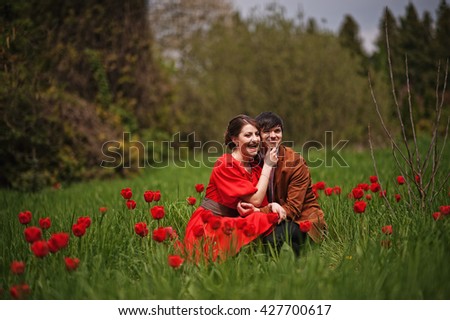 Couple hugging in love at red tulip field. Stylish man at velvet jacket and girl in red dress in love together