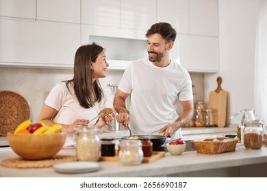 A couple is joyfully interacting while cooking in a modern kitchen. They are sharing smiles and laughter, preparing a healthy breakfast surrounded by fresh ingredients.