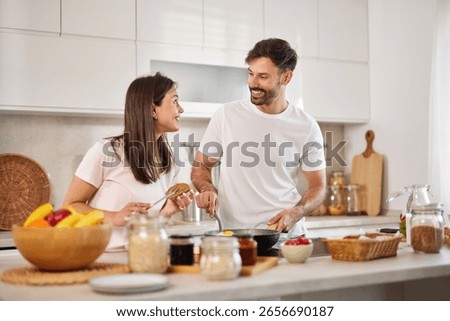 A couple is joyfully interacting while cooking in a modern kitchen. They are sharing smiles and laughter, preparing a healthy breakfast surrounded by fresh ingredients.