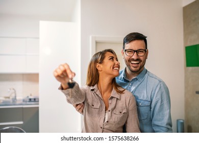 Couple With Keys To New Home. Cheerful Young Couple Holding Keys And Smiling While Standing In Their New House. Happy Couple In Their New Home Holding Key