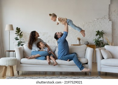 Couple with kids gathered in living room play together have fun sit on sofa, loving dad lifts on arms cute daughter, mom with son embracing resting on couch. Well-being family weekend at home concept - Powered by Shutterstock