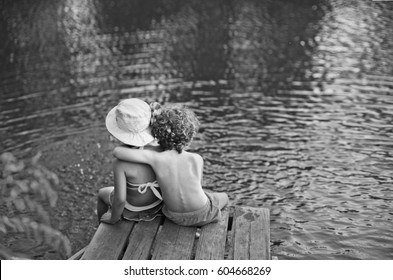 Couple Of Kids Seating On The Beach, Child Whispering On Wooden Platform, Summer Spring Sunny Morning, Jetty On The Water Lake Background, Vintage Black And White, The Boy Hugs The Girl From The Back