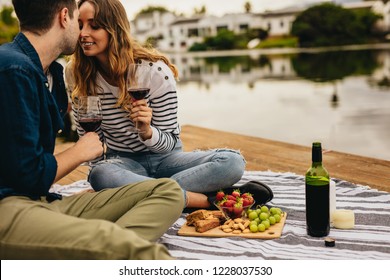 Couple Kissing Each Other On A Date Sitting Beside A Lake. Couple In Love Sitting On A Wooden Dock Near A Lake Drinking Wine And Snacks.