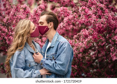 Couple kissing with protective masks. Love during quarantine of coronavirus pandemic conception. Man and woman posing near spring blossom pink trees. Copy, empty space for text