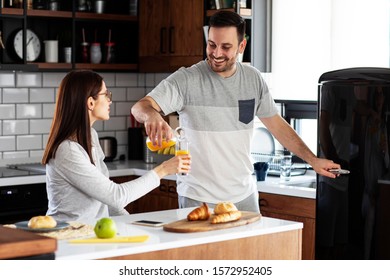 Couple In The Kitchen Eat Breakfast With Orange Juice And Pastry At Table And He Open Refrigerator