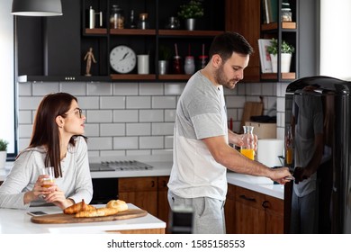 Couple In The Kitchen Eat Breakfast With Orange Juice And Pastry At Table And He Open Refrigerator