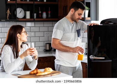 Couple In The Kitchen Eat Breakfast With Orange Juice And Pastry At Table And He Open Refrigerator