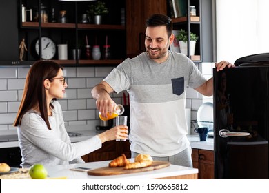 Couple In The Kitchen Eat Breakfast With Orange Juice And Pastry At Table And He Open Refrigerator