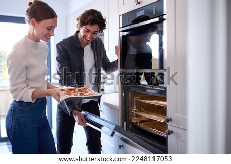 Couple In Kitchen At Home Putting Homemade Pizza Into Oven To Bake