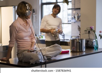 Couple In Kitchen With Woman Listening To Headphones And Man Drying Dishes