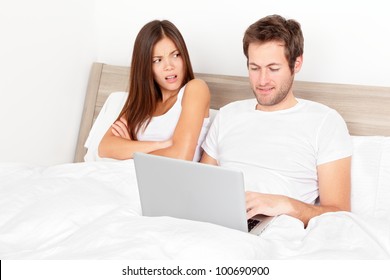 Couple With Laptop In Bed. Man At Computer, Woman Upset And Angry Looking At Man. Young Modern Interracial Couple In Bed. Asian Woman, Caucasian Man. Funny Image.