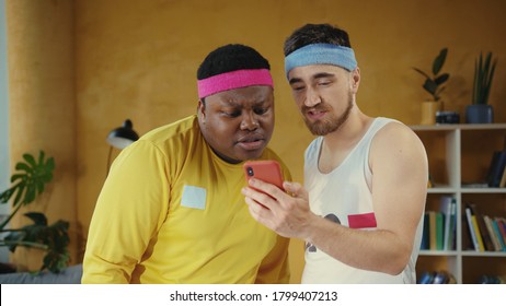 Couple Of Lazy Fitness Beginners Multi-ethnic Men Using Smartphones Watching Internet Content Working Out Together At Home. Fun, People, Technology.