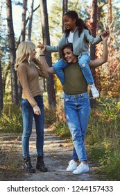 A Couple Of Lesbian Ladies With Their Adopted Teenage Daughter In The Autumn Park. The Brunette Girl Holding The Kid On Her Shoulders. The Young Family Standing, Holding Hands, Chatting, Smiling.