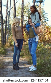 A Couple Of Lesbian Ladies With Their Adopted Teenage Daughter. The Brunette Girl Holding The Kid On Her Shoulders. The Young Family Standing In The Autumn Park, Holding Hands, Chatting, Smiling.