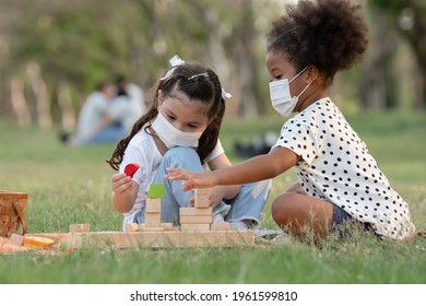 Couple Little Girls African And Caucasian Kids Wear Face Mask While Sitting And Playing Wooden Blocks Toy In Green Park Together. Ethnic Diversity Of Friendship In New Normal Lifestyle Concept