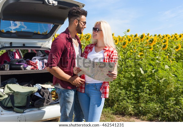 Couple looking road map standing sunflowers field\
outdoor, mix race man and woman standing near car trunk summer day\
holiday trip