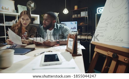  couple looking through documents at workplace. Focused african american man and beautiful woman reading business reports in office. Serious hipster team working with data indoors.