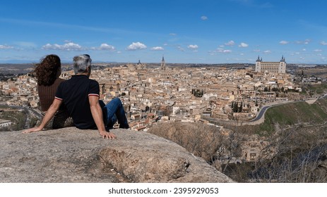 A couple looks out over the historic city of Toledo, a UNESCO World Heritage Site, sitting on the Piedra del Rey Moro, a well-known viewpoint over the city on a sunny day. Tourism concept in Spain - Powered by Shutterstock