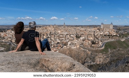 A couple looks out over the historic city of Toledo, a UNESCO World Heritage Site, sitting on the Piedra del Rey Moro, a well-known viewpoint over the city on a sunny day. Tourism concept in Spain