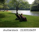 A couple lounges in Adirondack chairs in the midst of a green lawn on the shores of a still lake at Peaks of Otter Lodge along the Blue Ridge Parkway in Virginia.