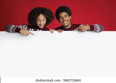 Couple In Love Advices Great Proposal For New Year And Christmas. Happy Young African American Family Looking At Blank White Billboard And Pointing With Fingers At Banner, Isolated On Red Background