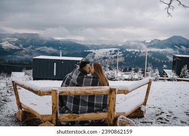 Couple in love with blanket hugs, kisses and admiring winter mountains. Recreation area modern house, fire pit, fireplace in backyard. Cabin terrace with mountain view. Tiny house on a hill. Back view - Powered by Shutterstock