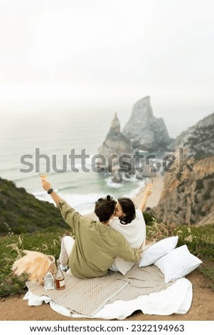 Couple in love celebrating their engagement on the seashore, drinking champagne with breathtaking view at coastline, copy space. Honeymoon trip, wedding travel. Vertical shot