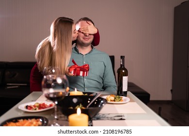 Couple In Love Celebrating Valentine's Day At Home, Girl Gives A Gift To A Man, Kisses Him With Her Hand Covering Her Eyes, Date Of Lovers At Dinner, Gift Concept For Valentine's Day Or Anniversary