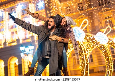 Couple in love having fun celebrating New Year outdoors in city streets or square, boyfriend piggybacking girlfriend with Christmas lights in background - Powered by Shutterstock