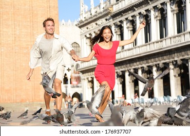 Couple In Love Having Playful Fun In Venice Holding Hands Running Laughing In Venice, Italy On Piazza San Marco. Happy Young Couple On Travel Vacation On St Mark's Square. Happy Woman And Man.