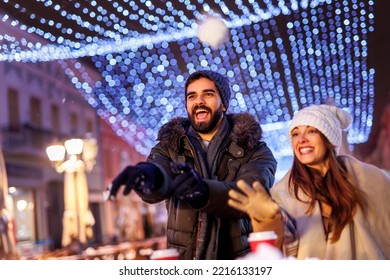 Couple In Love Having A Snowball Fight While Spending Christmas Eve Outdoors In The Nicely Decorated City Streets With Christmas Lights All Around