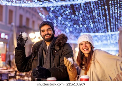 Couple In Love Having A Snowball Fight While Spending Christmas Eve Outdoors In The Nicely Decorated City Streets With Christmas Lights All Around