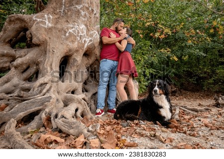 A couple in love hugging and kissing under a tree with large crisscrossed and prominent roots, while their  Bernese mountain dog is keeping a watch.