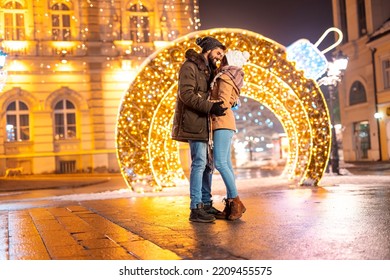 Couple in love hugging on city square with Christmas lights all around, spending time outdoors in the nicely decorated city streets during winter holiday season and advent
