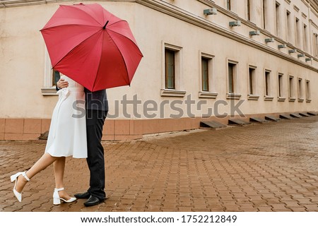 Couple in love kissing under red umbrella. Cute couple standing under umbrella. They closed with an umbrella from people