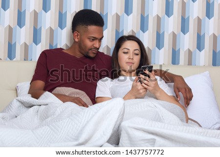 Couple in love lieing on white bed. Young darkskinned man and woman looking at mobile phone, reading or watching something interesting, handsome man hugging her wife, spending time together.