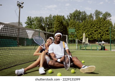 Couple in love likes to play tennis. After game, they sat down on grass of tennis court, hugging, smiling and enjoying the moment. Young people hold rackets. Yellow tennis balls lie on the ground. - Powered by Shutterstock