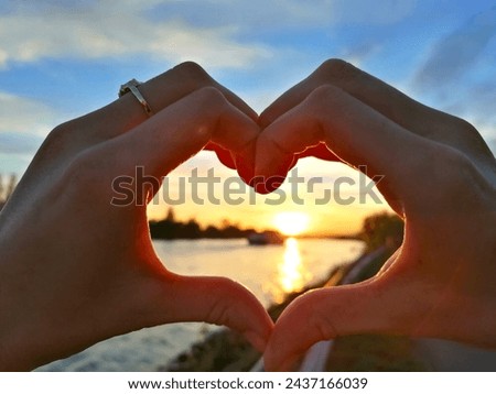 A couple in love makes a heart with their hands against the backdrop of a sunset by the river