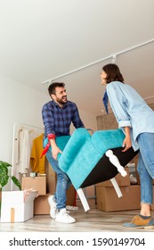 Couple In Love Moving In Together, Having Fun While Carrying An Armchair And Setting Up The New Furniture Arrangement