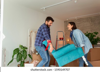 Couple In Love Moving In Together, Having Fun While Carrying An Armchair And Setting Up The New Furniture Arrangement