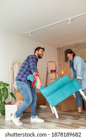 Couple In Love Moving In Together, Having Fun While Carrying An Armchair And Setting Up The New Furniture Arrangement