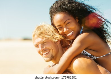 Couple in love on beach - Caucasian man having his Hispanic woman piggyback on his back under blue sky near ocean