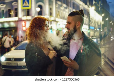 Couple in love on date. Boyfriend holds phone. She hugs him and smokes an electronic cigarette. They look at each other in street of night city. Wet asphalt after rain.