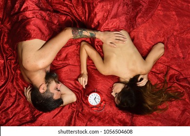 Couple in love on red sheets. Sex and perfect morning concept. Man and woman with half covered bodies lie near red alarm clock. Guy with beard and tattoos touches pretty ladys back in bed, top view