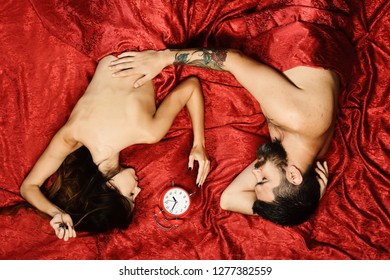 Couple in love on red sheets. Man and woman with half covered bodies lie near red alarm clock. Guy with beard and tattoos touches pretty ladys back in bed, top view. Love and perfect morning concept.